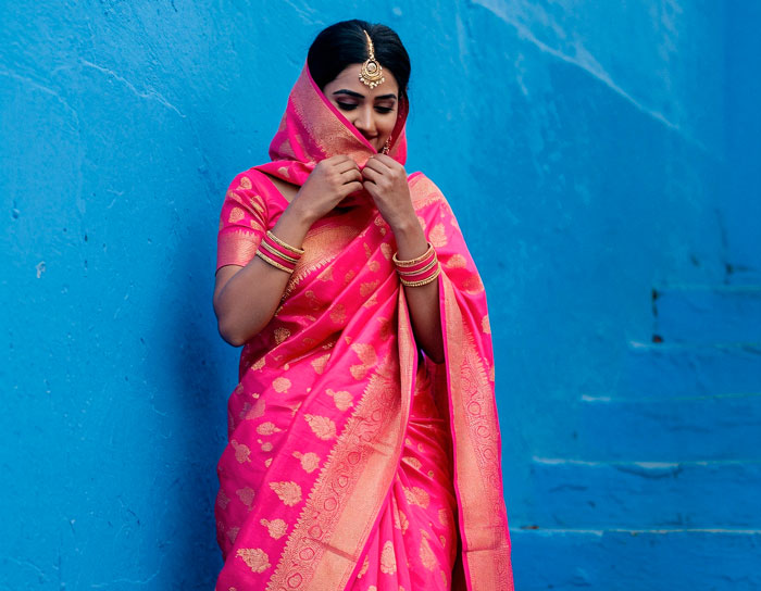 Woman in traditional pink sari adjusting her attire against blue wall, highlighting cultural attire and cultural appropriation debate.