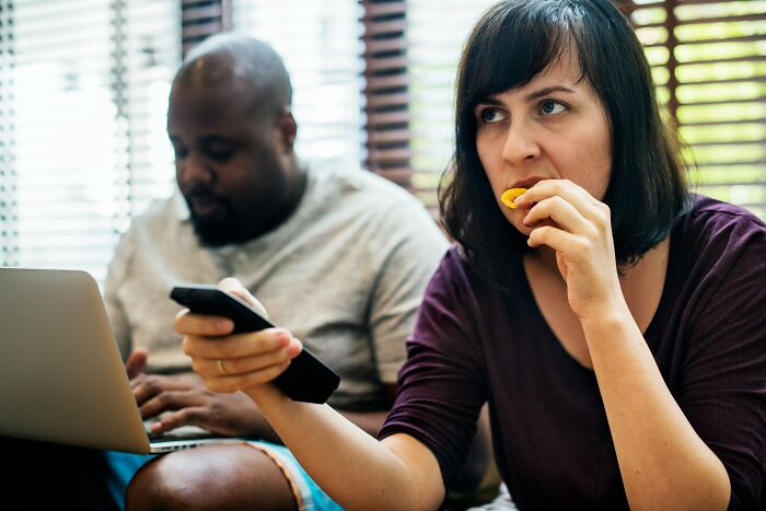 A woman holding a remote and snacking while a man uses a laptop, illustrating ways to avoid babysitting.