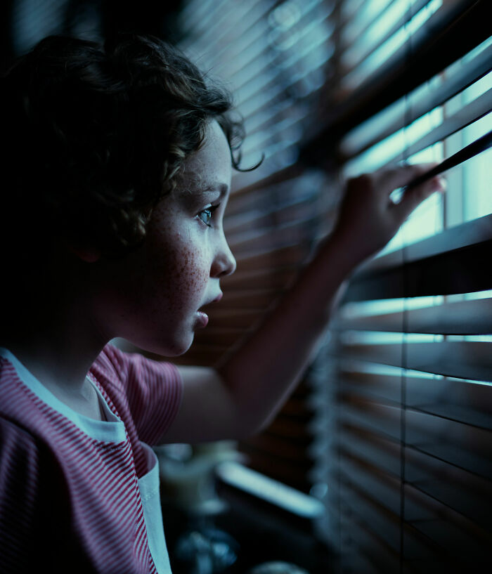 Child with freckles peering through window blinds at night, evoking unsettling and terrifying things people swear they've seen.