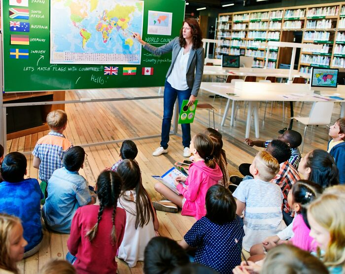 Teacher pointing at world map on blackboard while diverse children sit on floor in modern classroom setting about global studies and modern life.