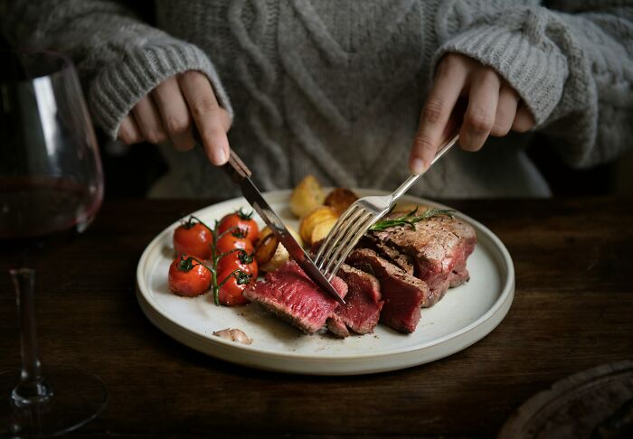 Person in knit sweater cutting rare steak with roasted tomatoes on plate, illustrating wealth gap perspective concept.