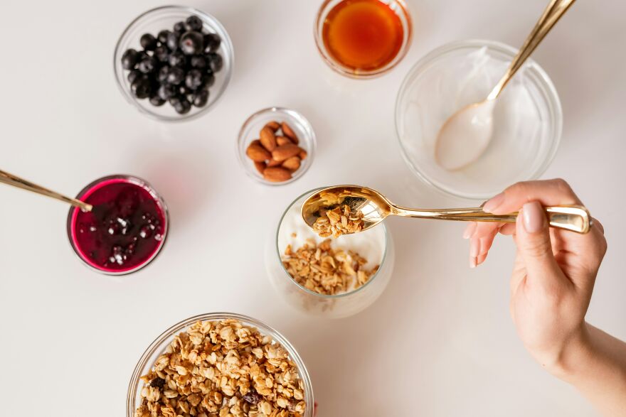 Hand holding a gold spoon scooping granola with yogurt, blueberries, almonds, and honey on a white table.
