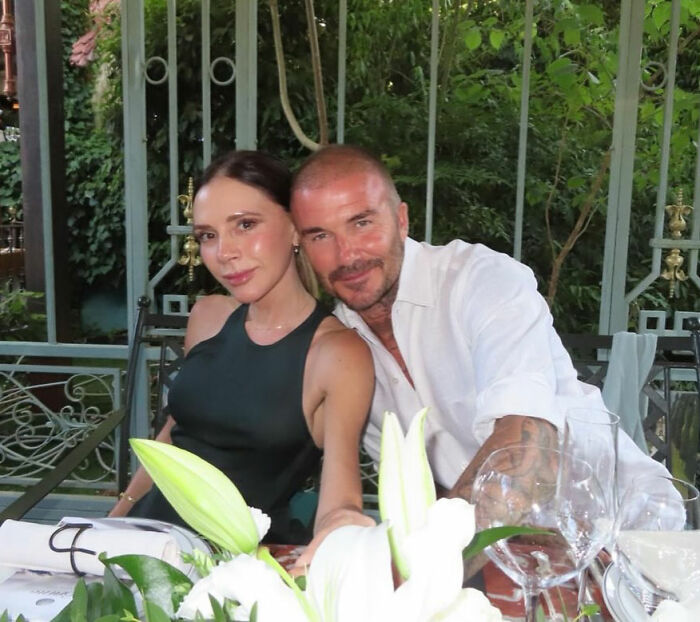 David and Victoria Beckham sitting together at an outdoor table amid greenery, looking heartbroken.