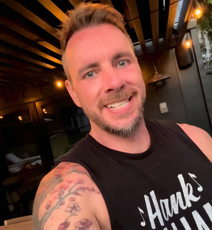 Dax Shepard smiling in a black tank top with a floral tattoo on his arm in a warmly lit indoor setting.