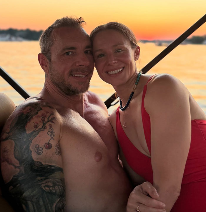 Dax Shepard smiling shirtless next to Heidi Klum in a red dress enjoying a sunset moment on the water.