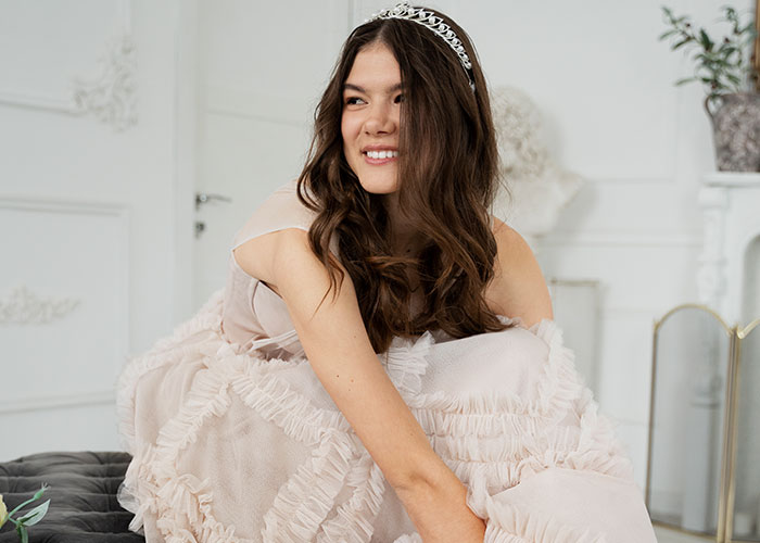 Young woman wearing a tiara and a ruffled dress smiling softly, reflecting on disgusting things discovered about friends.
