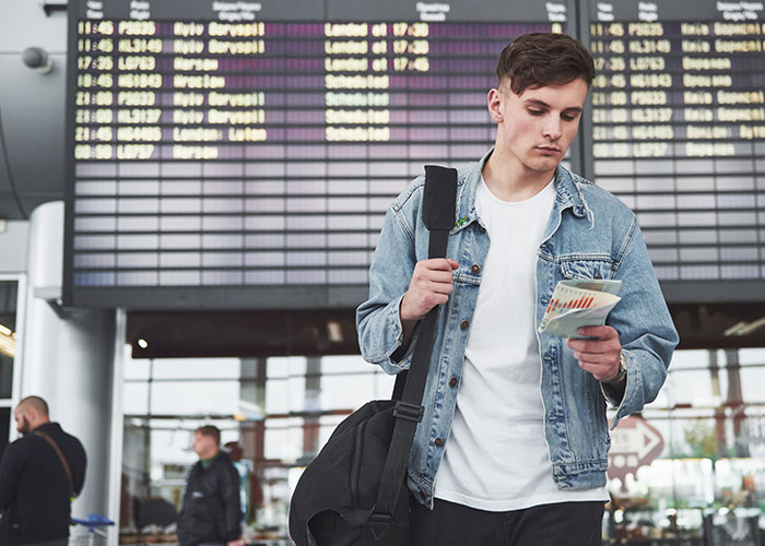 Young man at airport looking at boarding pass with flight schedule in background, reflecting on disgusting things about friends.