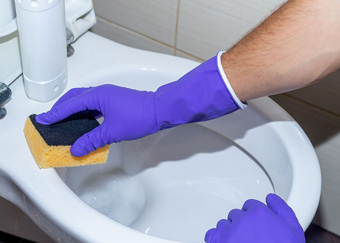 Person wearing purple gloves cleaning a toilet with a sponge, highlighting disgusting things people discovered about friends.