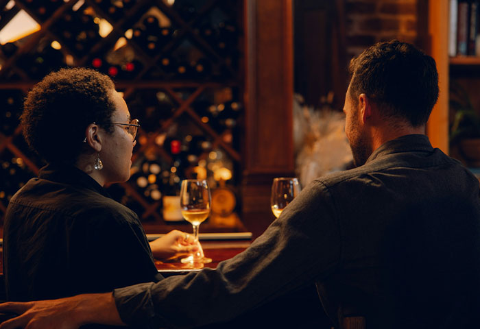 Man and woman sitting at wine bar having a serious conversation about divorce, with warm lighting and shelves in background.