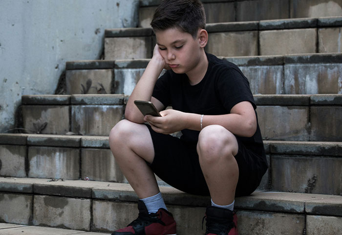 Boy in black outfit sitting on outdoor stairs, looking at phone with a sad expression about dad lies and divorce truth.