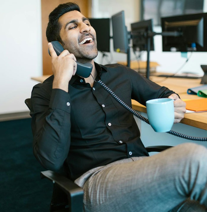 Man on phone laughing while holding a coffee mug, dealing with calls meant for a doctor in office setting.