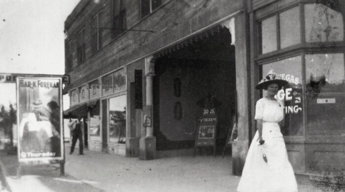 Historic black and white photo of early Vegas street scene showing vintage buildings and a woman in period clothing.
