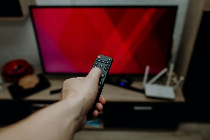 Hand holding a remote control pointing at a TV screen, symbolizing heartwarming and harmless secrets shared quietly.