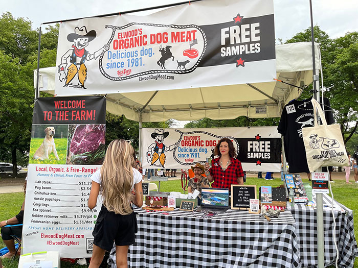 Outdoor stall promoting Elwood's organic dog meat with free samples, featuring banners and a woman behind the counter - 12