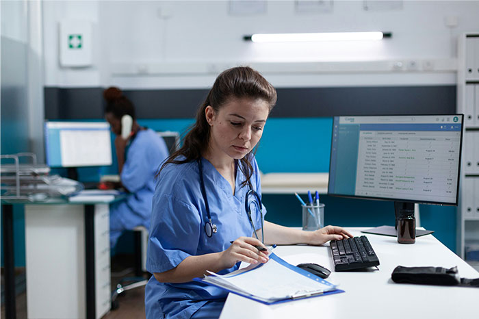 Female nurse in blue scrubs reviewing patient records on a clipboard in an emergency room setting with medical data on screen.