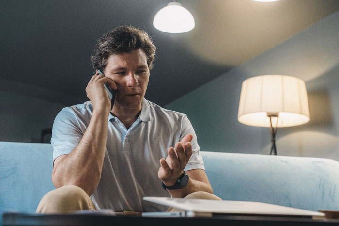 Man on phone looking concerned while sitting on couch at home, illustrating couple hosting old friend regrets. Man on phone looking concerned while sitting on couch at home, illustrating couple hosting old friend regrets.