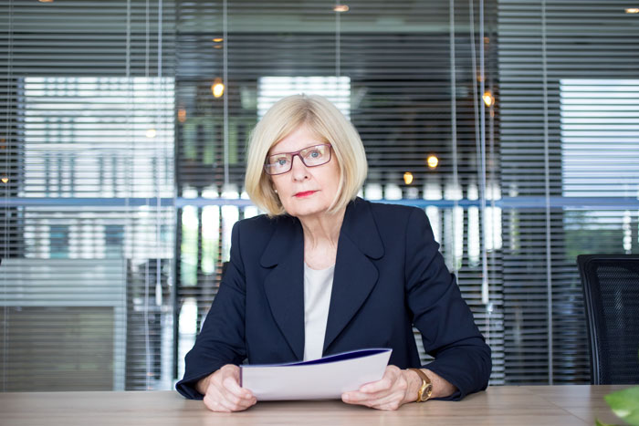 Older woman in business attire sitting at desk holding papers, representing coworker retirement party and forced quit scenario