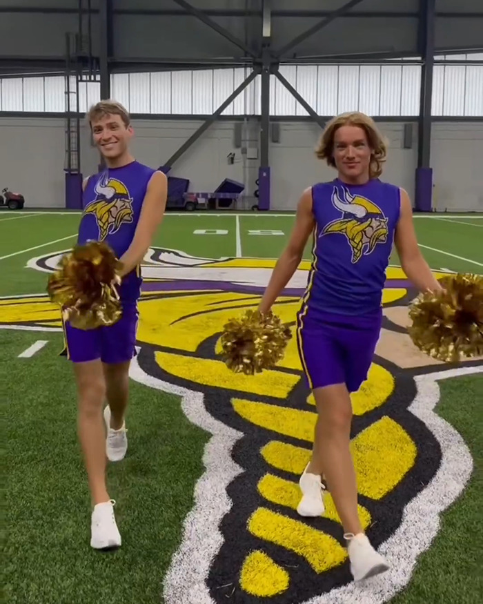 Two male cheerleaders in purple uniforms perform with gold pom-poms on an American football field indoors.