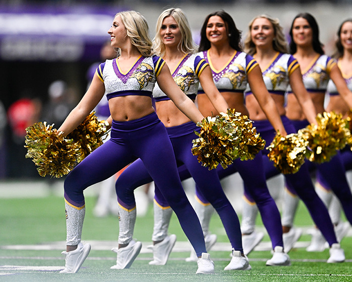 Cheerleaders performing on football field in purple and gold uniforms with pom-poms during a halftime routine.