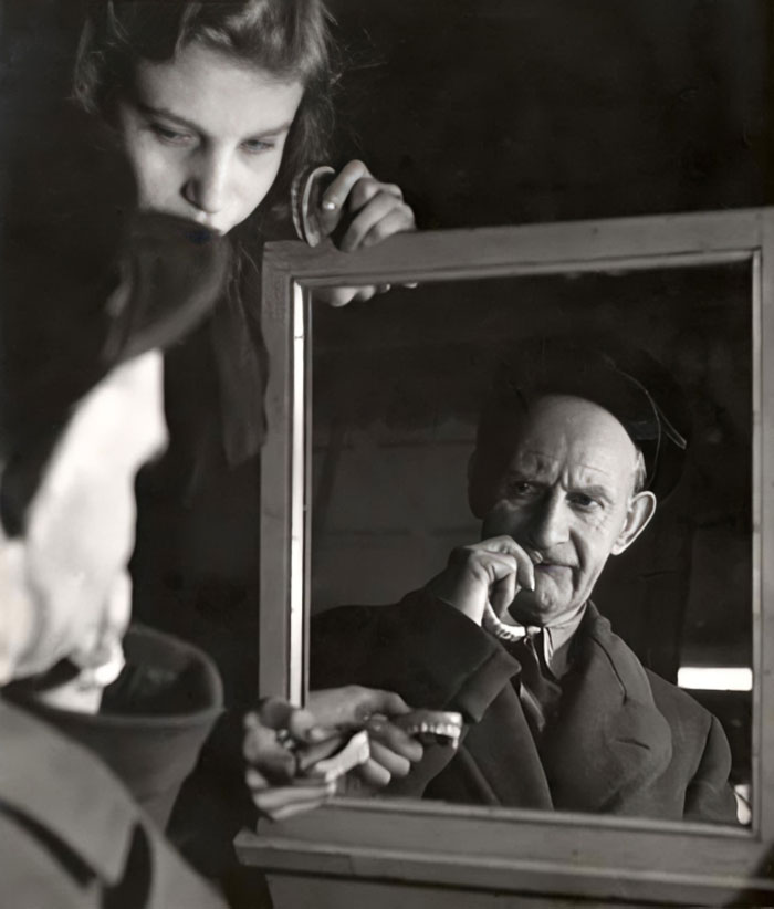 Man brushing his teeth reflected in a mirror held by a woman in a vintage black and white historical photo.