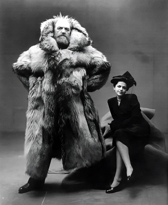 Black and white historical photo of a man in a large fur coat standing next to a seated woman in a hat and dress