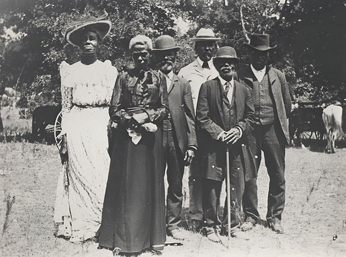 Group of six people dressed in early 20th century clothing posing outdoors in a rare historical photo.