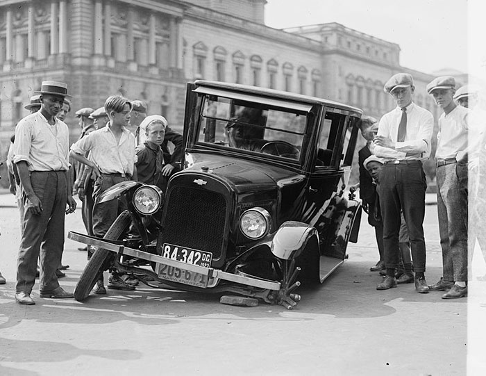 Vintage historical photo of a car stuck on a bicycle wheel with a crowd of onlookers in an urban setting.