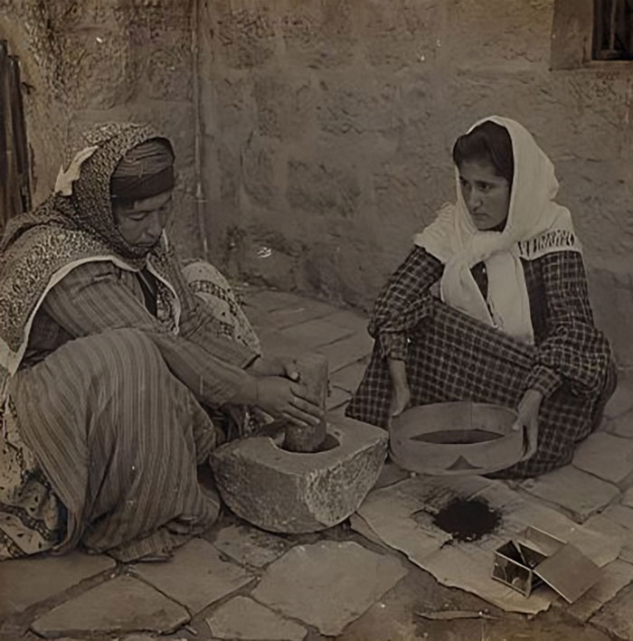 Two women in traditional clothing preparing food using a stone grinder in a historical photo with vintage atmosphere