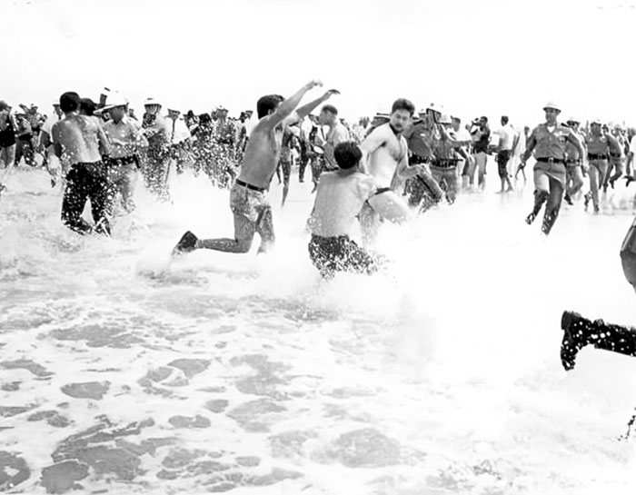 Black and white historical photo showing a chaotic beach scene with shirtless men and uniformed officers splashing in the water.