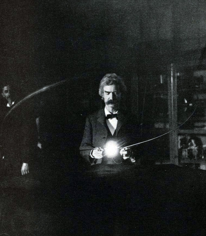 Black and white historical photo of a man demonstrating an early light bulb in a dark room with visible wiring.
