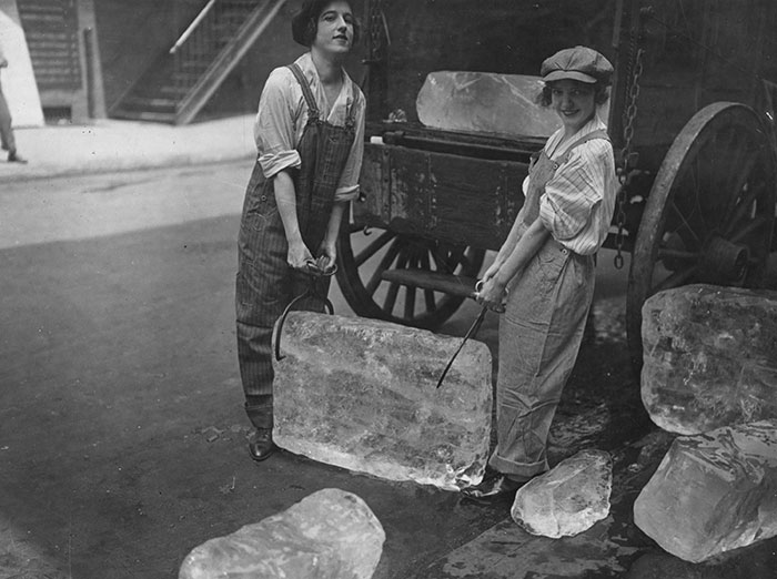 Two women in vintage work clothes lifting a large ice block on a street, a rare historical photo with unique charm.
