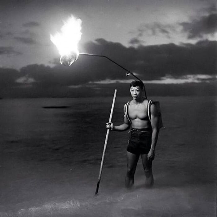 Historical photo of a man standing in water holding a flaming torch attached to his back at night.