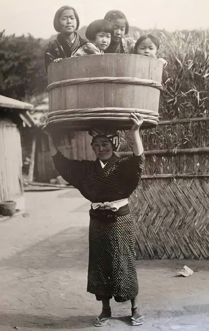 Woman carrying a large wooden bucket on her head with four children inside in a historical black and white photo.