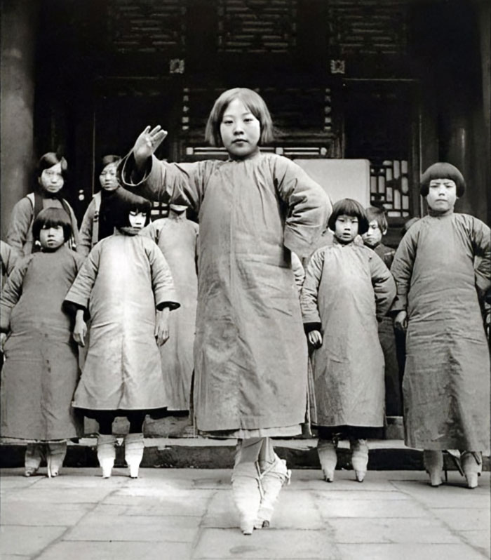 Group of children wearing traditional clothes and balancing on tall wooden stilts in a historical photo.