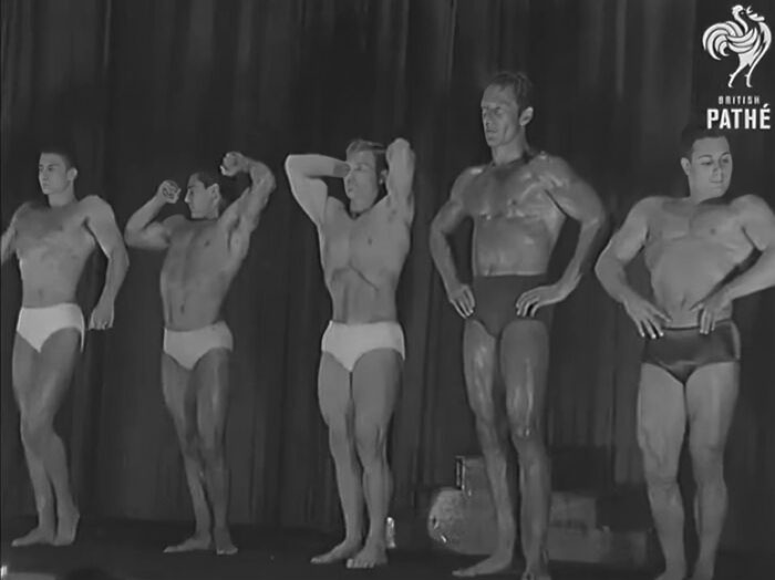 Black and white historical photo of five bodybuilders striking poses on stage, showcasing unusual muscular forms.