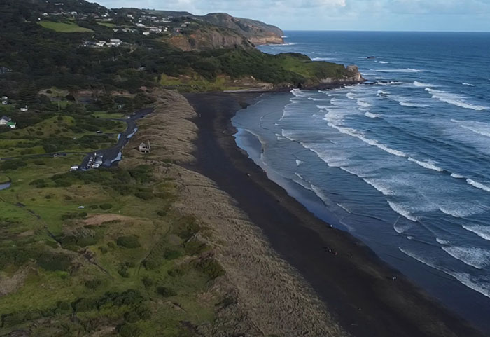 Aerial view of a black sand beach and ocean waves near green cliffs under a partly cloudy sky.