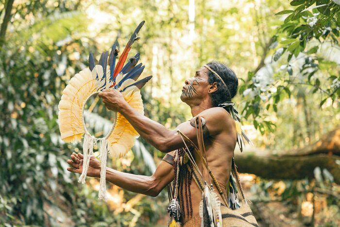 Indigenous man in traditional attire holding feathered headdress in forest, symbolizing good news and cultural heritage spotlight.