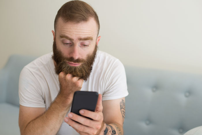 Bearded man looking concerned at his phone while sitting indoors, depicting a fiance destitute relationship moment. Bearded man looking concerned at his phone while sitting indoors, depicting a fiance destitute relationship moment.