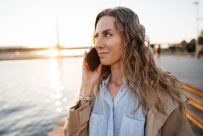 Woman with long wavy hair talking on phone by waterfront during sunset, reflecting on a fiance destitute relationship. Woman with long wavy hair talking on phone by waterfront during sunset, reflecting on a fiance destitute relationship.