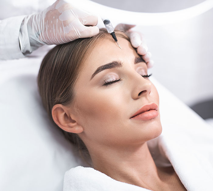 Young woman receiving cosmetic filler injection on forehead from gloved professional in a clinical setting related to Love Island face trend.