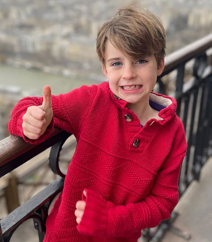 Young boy in a red sweater giving thumbs up outdoors near a metal railing, related to Minneapolis school attacker debate.