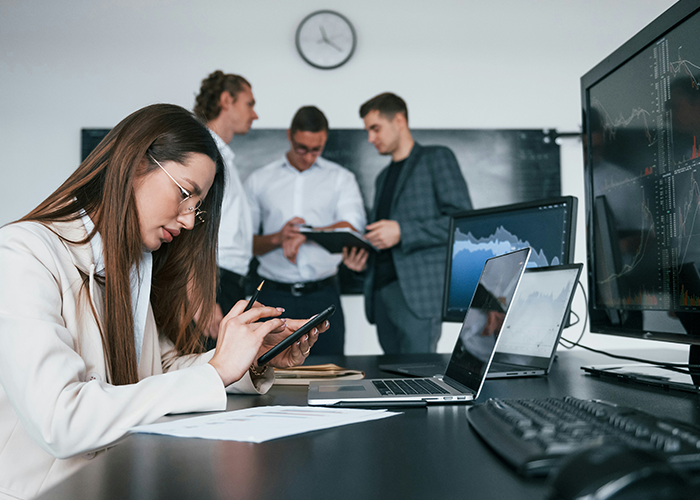Woman working on tablet in office setting while colleagues discuss, illustrating issues with boss banning flexible hours at work. Woman working on tablet in office setting while colleagues discuss, illustrating issues with boss banning flexible hours at work.