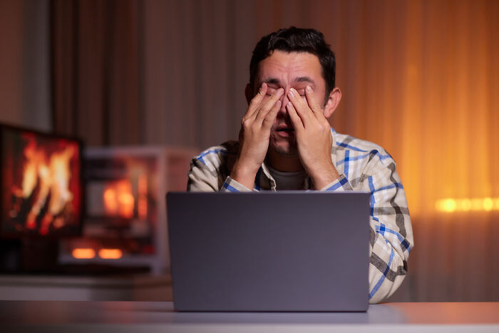 Man in plaid shirt reacting with shock and rubbing eyes while viewing creepy things on Reddit screen at night.