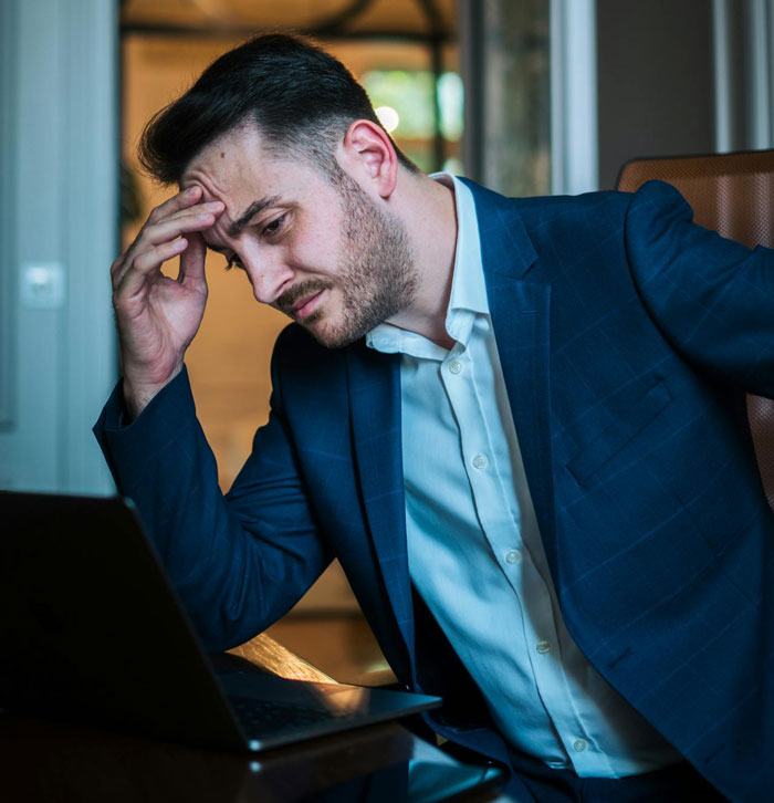 Stressed man in business attire at laptop, depicting effects of boss going out of his way to make life difficult for employee.