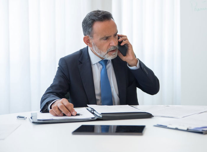 Middle-aged businessman in a suit and tie looking concerned while speaking on the phone at his office desk