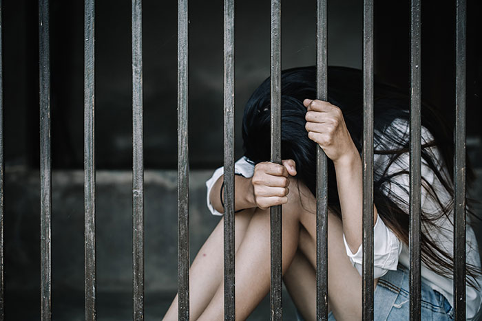A young inmate sitting behind prison bars, clutching the metal bars, illustrating life in prison conditions.