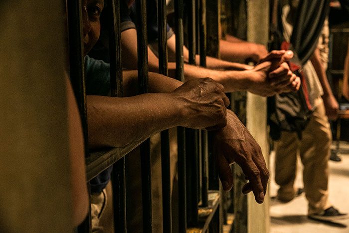 Prison inmates reach hands through bars in a dimly lit cell, illustrating life in prison conditions.