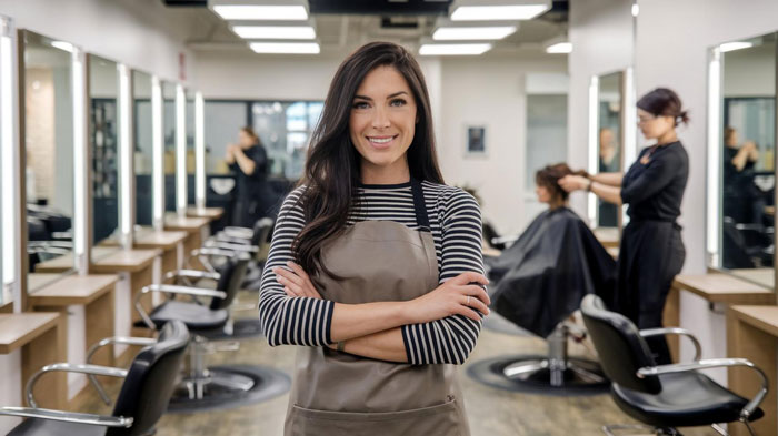 Young female stylist in a free salon service setting smiling confidently with clients getting hair treatments in the background Young female stylist in a free salon service setting smiling confidently with clients getting hair treatments in the background