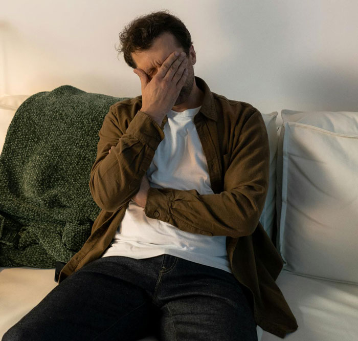 Man sitting on couch with hand covering face, stressed about friend breaking into house and planting belongings.