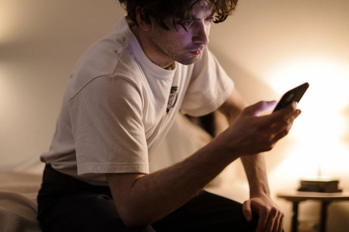 Young man sitting on bed, looking at phone with focused expression in a dimly lit room, illustrating friend breaks into house scenario.